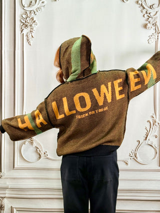 A person shown from behind wearing a brown knit hoodie with 'HALLOWEEN' and 'TRICK OR TREAT' printed in yellow across the back, featuring green and yellow striped hood and cuffs, paired with black pants, set against an ornate white wall.