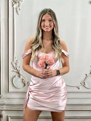 A woman with blonde-highlighted hair stands indoors wearing a light pink satin off-the-shoulder mini dress and holding a bouquet of pink roses. The background is a white decorative wall with ornate molding.