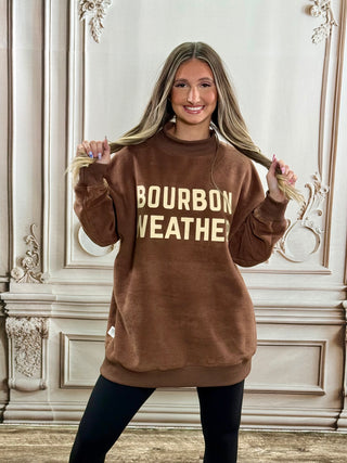 A woman modeling a brown sweatshirt with 'BOURBON WEATHER' printed in gold lettering, posing in front of an ornate cream-colored wall.