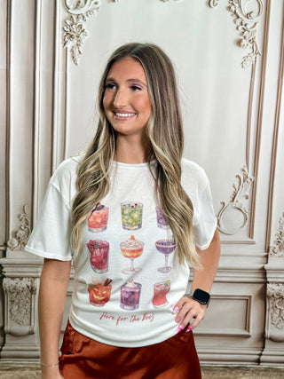 A woman with long blonde-highlighted hair stands indoors wearing a white graphic T-shirt with colorful cocktail illustrations and the text 'Here for the bars,' paired with rust-colored satin pants. The background is an ornate cream-colored wall with decorative molding.