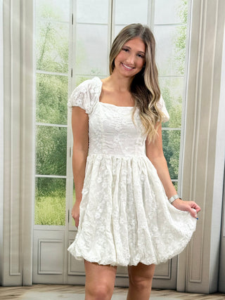 Woman wearing a white lace dress standing in a room with large windows.