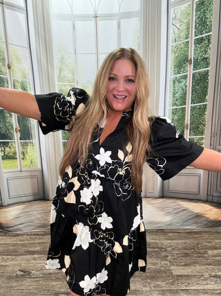 Woman wearing a black floral dress standing indoors with large windows in the background.