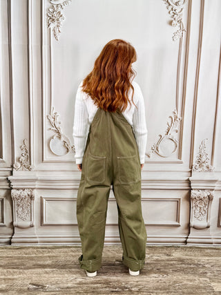 A woman with long red hair is shown from behind wearing olive green wide-leg overalls over a white ribbed long-sleeve top, standing in front of an ornate light gray wall.