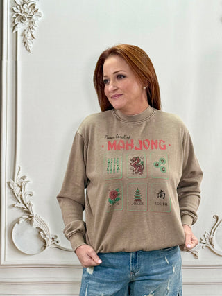 A woman with red hair wearing a beige crewneck sweatshirt with a Mahjong graphic print, paired with blue jeans, standing against a white wall with ornate molding details.
