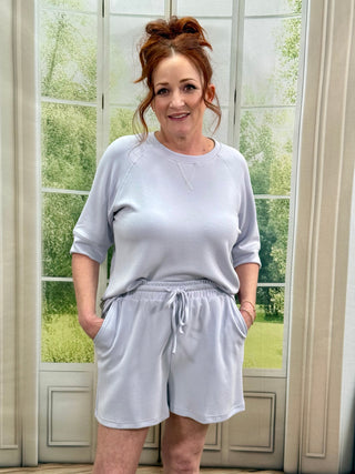 Woman wearing a light blue sweatshirt and shorts standing in front of a window with greenery outside.