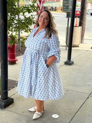 Woman in a blue patterned dress standing on a sidewalk with trees and street elements in the background.