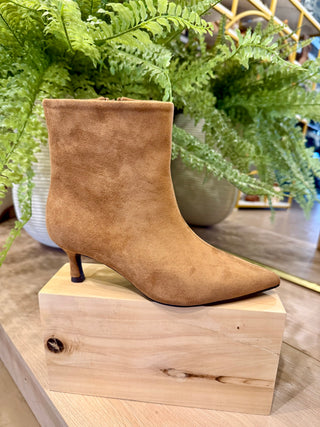 A tan suede ankle boot with a pointed toe and kitten heel, displayed on a light wood block with green ferns in the background.