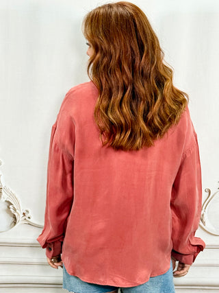 A woman with long wavy auburn hair is shown from behind wearing a dusty rose long-sleeve linen shirt and blue jeans, standing against a white wall with decorative molding.