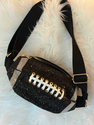 A black glitter football-shaped fanny pack with white laces, gold eyelets, and silver rhinestone stripes, featuring a black adjustable strap with a gold buckle, displayed on a white faux fur background.