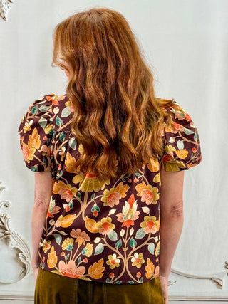 A woman viewed from behind wearing a short-sleeve blouse with a brown base and a colorful floral pattern in orange, yellow, and green, paired with olive green pants.