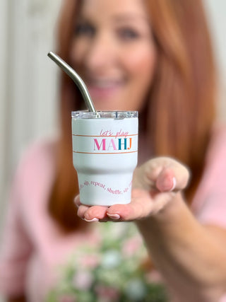 A woman holds up a white insulated tumbler with a metal straw. The tumbler has pink and blue text that reads 'let's play MAHJ' with smaller pink text below.