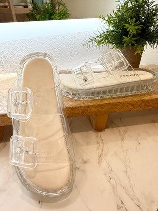 Beige slide sandals with clear straps on a marble surface with a plant in the background