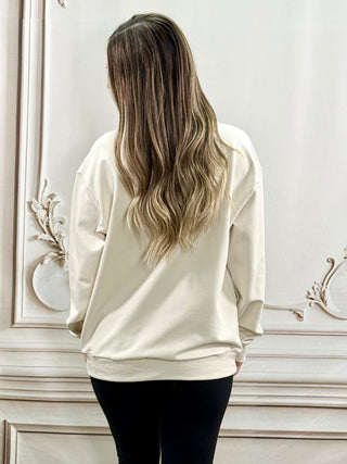 A woman with long, wavy blonde-highlighted hair is shown from behind, wearing an oversized cream sweatshirt and black pants, standing in front of a white wall with decorative molding.