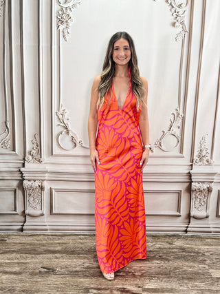 A woman wearing a sleeveless, floor-length halter dress with a bold orange and pink tropical leaf pattern, standing in front of an ornate light gray wall.