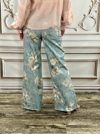 A person shown from the back wearing wide-leg light blue jeans with a faded floral print, paired with a pale pink blouse, standing in front of a decorative paneled wall.