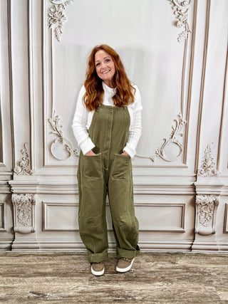 A woman with red hair stands in front of an ornate light gray wall, wearing olive green wide-leg bib overalls over a white long-sleeve top and brown sneakers.