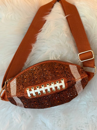 A football-shaped shoulder bag covered in brown glitter, featuring white laces, white and brown stripes, and a brown adjustable strap with a gold buckle, displayed on a white faux fur background.
