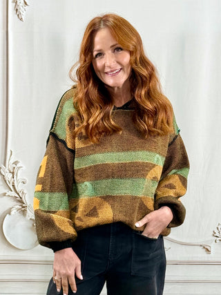 A woman with long red hair wearing a brown and green striped knit sweater and black pants, standing in front of a white decorative wall.