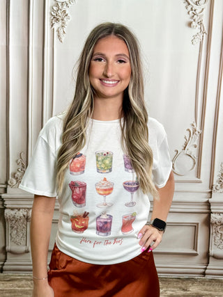 A smiling woman with long blonde-highlighted hair stands indoors wearing a white graphic T-shirt with colorful cocktail illustrations and the text 'Here for the Boys,' paired with rust-brown satin pants. The background is an ornate cream-colored wall with decorative molding.