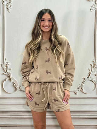 A woman modeling a matching beige lounge set with a brown poodle print, consisting of a long-sleeve sweatshirt and drawstring shorts, posed against an ornate white wall.