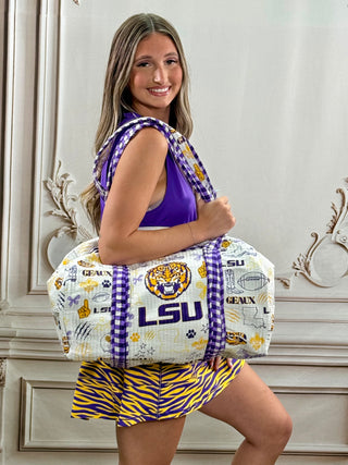 A woman wearing a purple and yellow LSU-themed outfit carries a large white duffel bag with purple and yellow plaid straps and a colorful LSU tiger logo print, posed in front of an ornate cream-colored wall.