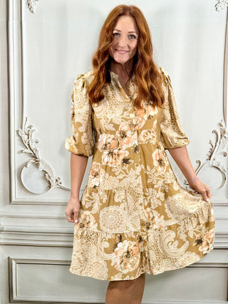 A woman with long red hair models a short-sleeved, button-front dress with a beige and cream floral paisley pattern, standing against a white paneled wall.