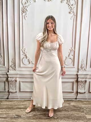 A woman modeling a floor-length cream satin gown with puff sleeves and a lace-trimmed bodice, standing in front of an ornate beige paneled wall.