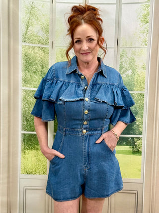 Woman wearing a blue denim romper standing in front of a window with greenery outside.