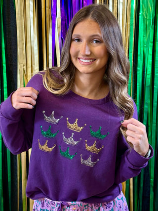 Woman wearing a purple sweatshirt with embroidered crowns in front of a colorful curtain.