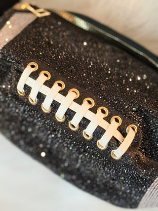 A close-up of a black glittery football-shaped pouch with gold laces and a gold zipper pull, shown against a light neutral background.