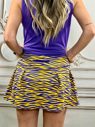 A woman shown from the back wearing a purple sleeveless top and a pleated mini skirt with a bold yellow and purple wavy pattern, standing indoors against a light-colored wall.