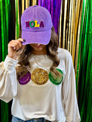 Person wearing a white long-sleeve shirt with colorful designs and a purple 'NOLA' cap against a metallic curtain background.