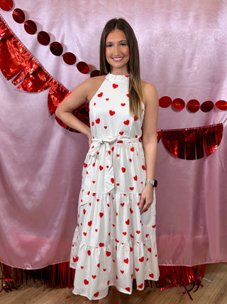 Woman in a white dress with red heart patterns standing in front of a decorative pink and red background.