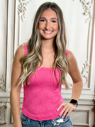 A smiling young woman with long blonde-highlighted hair wearing a bright pink textured tank top and blue jeans, standing indoors against an ornate cream-colored wall.