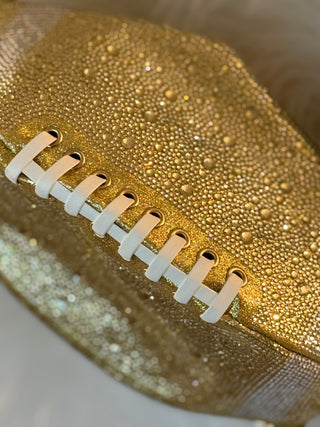 A close-up of a gold glittery football-shaped item with white laces, showing a sparkling textured surface and metallic finish.