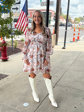 Woman in a floral dress and white boots standing on a sidewalk with an American flag in the background.