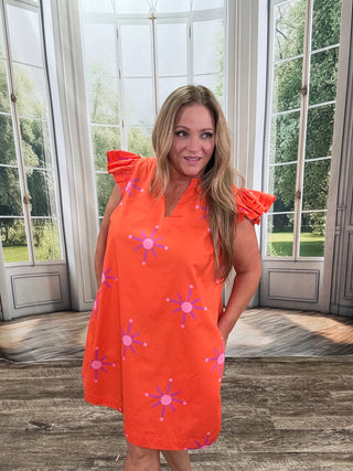 Woman wearing an orange dress with pink floral patterns in a room with large windows.