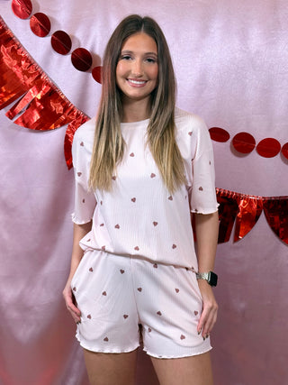 Woman in a white outfit with heart patterns standing against a decorative red and pink background.