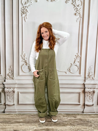 A woman with red hair stands in front of an ornate light gray wall, wearing olive green utility overalls over a white long-sleeve top and white sneakers.