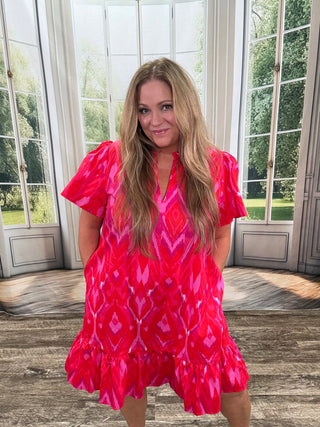 Woman wearing a red patterned dress standing indoors with a window in the background