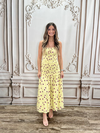 A woman modeling a yellow strapless maxi dress with brown polka dots, standing in front of an ornate beige wall with a rustic wooden floor.