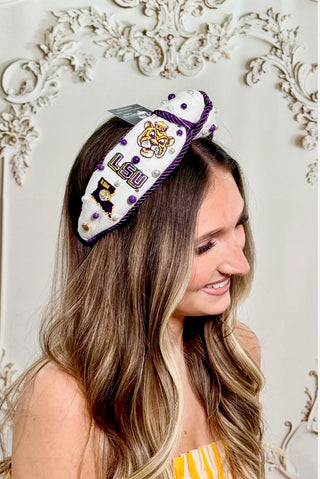 A woman with long highlighted brown hair wearing a white LSU Tigers themed headband with purple trim and gold accents, posed against a white ornate wall.