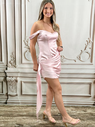 A woman models a light pink satin off-the-shoulder mini dress with a side tie detail, standing in front of an ornate white wall.