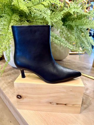 A single black ankle boot with a pointed toe and kitten heel, displayed on a light wood block with green ferns in the background.