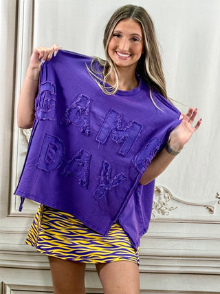 A woman holds up a purple oversized sweatshirt with 'GAME DAY' embroidered in 3D letters. She is wearing a yellow and purple striped skirt, standing against a light-colored paneled wall.