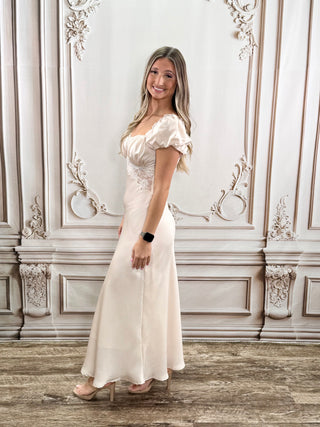 A woman modeling a floor-length cream or ivory evening gown with puffed short sleeves, a sweetheart neckline, and a fitted skirt, standing in front of an ornate white paneled wall.