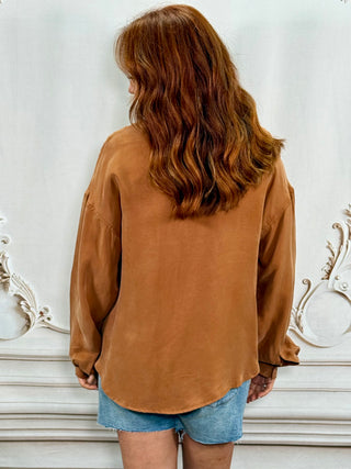 A woman with long wavy auburn hair is shown from behind, wearing an oversized brown long-sleeve top and denim shorts against a white decorative wall.