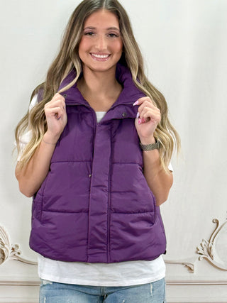 A woman wearing a purple quilted puffer vest over a white t-shirt and blue jeans, standing in front of a light-colored wall with decorative molding.