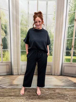 Person wearing a black outfit standing in front of a large window with a view of greenery.