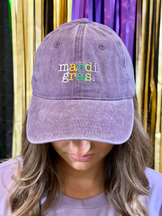 Purple cap with 'mardi gras.' text worn by a person against a colorful background
